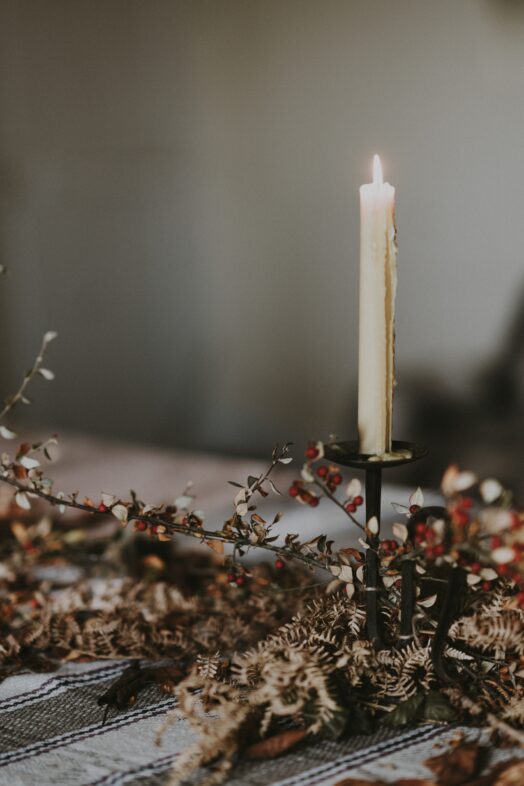 Ett ensamt tänt ljus står i en ljusstake, omgiven av torkade blommor på ett bord.